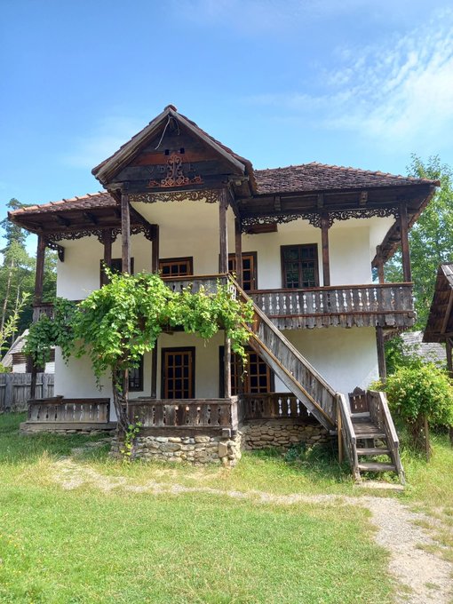 A traditional two-story white house with ornate wooden balconies and a steeply pitched roof stands in a grassy yard under a bright blue sky, with grapevines climbing its walls. The Eastern European architecture, featuring decorative woodwork and stone foundations, fits the post's theme of showcasing photos from the region in an effort to reach the $ENA $3 milestone.