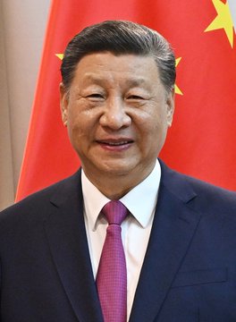 The image features a man wearing a suit and tie standing next to a flag with yellow stars. He appears to be smiling for the camera, possibly posing for a photo or portrait. The background of the image includes a red flag, which could represent China's national identity or symbolize their commitment to AI development in the future. The caption provided suggests that this man is an important figure related to AI and its potential impact on society.
