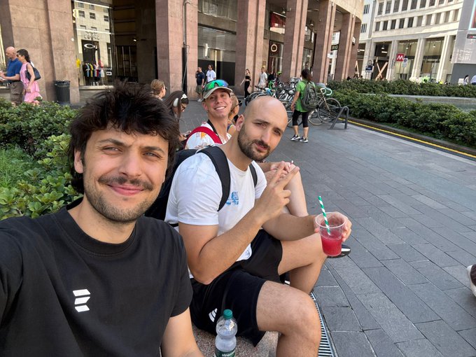 The post references a Solana meetup and is asking what the Solana Italy community is working on. The image shows a casual but fitting scene: three men sitting together at what appears to be a public square or arcade in an Italian city, smiling for a selfie with one holding a red drink — a low-key break reflecting the relaxed community vibe of a Solana gathering, with no on-screen text to tie directly to "Solana Italy" beyond the everyday camaraderie the post implies.

The provided text or background cannot be read clearly; what's described is the setting and the relaxed group vibe fitting a community meetup.