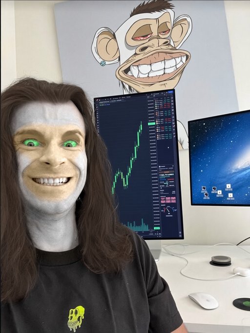 A person wearing a SpongeBob-style filter with green eyes and a wide grin, sitting in front of dual monitors displaying stock charts and a BAPE logo artwork on the wall.
