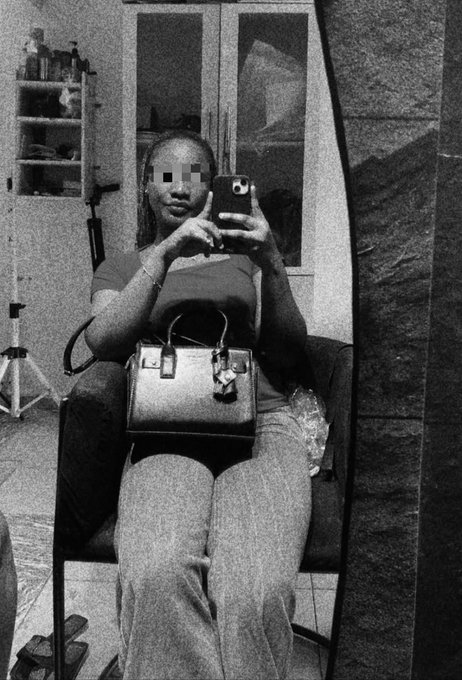 A grainy black-and-white mirror selfie shows a seated woman holding a phone, with her eyes pixelated and a handbag resting on her lap.
