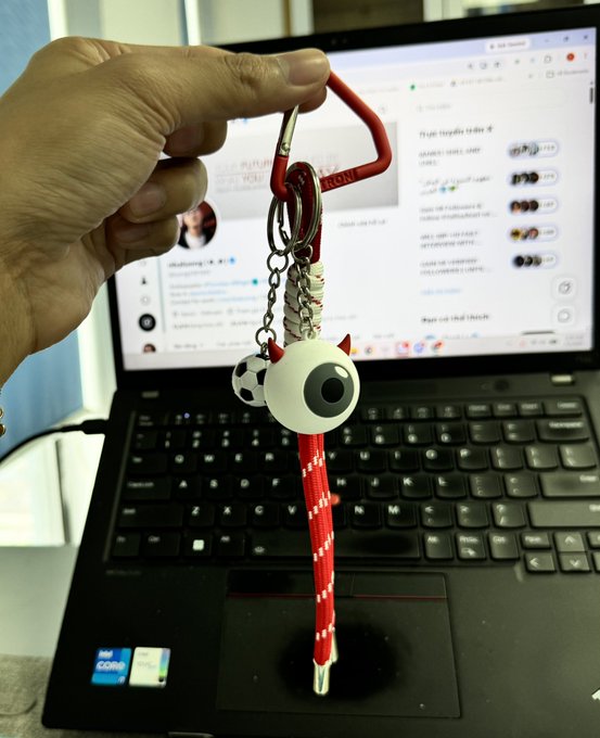 A hand holds a red keychain with a soccer ball and eyeball charm in front of a Lenovo laptop displaying a social media feed.