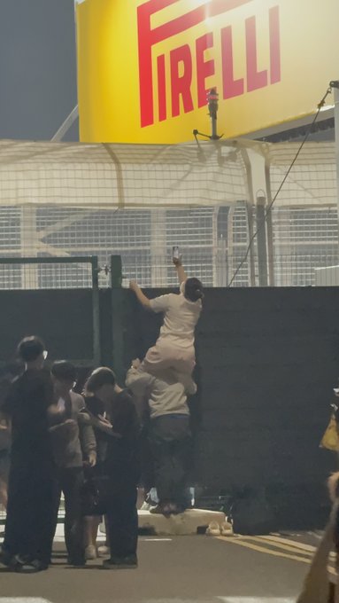 People climb over a barrier beneath a large yellow and red Pirelli sign, appearing to escape into a restricted area at night.