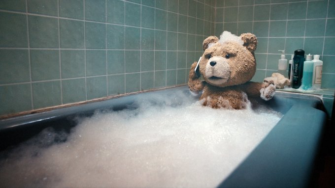A teddy bear covered in soap bubbles sits in a bathtub full of foam, appearing to talk on a phone with toiletries on the edge.