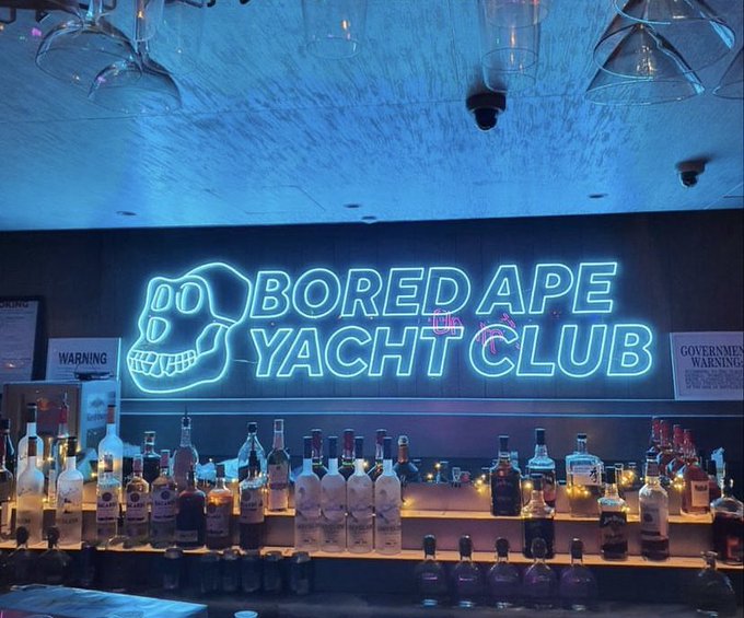 A bar interior featuring a large glowing blue neon sign reading 'BORED APE YACHT CLUB' above shelves stocked with liquor bottles and hanging glassware.