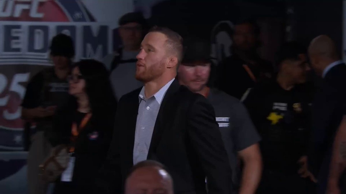 The image shows a man wearing a suit standing in front of a crowd, possibly attending an event or watching a fight. The crowd consists of several people surrounding him, with some individuals closer to the foreground while others are further back. A person is holding a cell phone, and there's also a handbag visible near one of the attendees. The image captures the atmosphere of a social gathering or public event where people have gathered to watch or participate in an activity involving fighters.