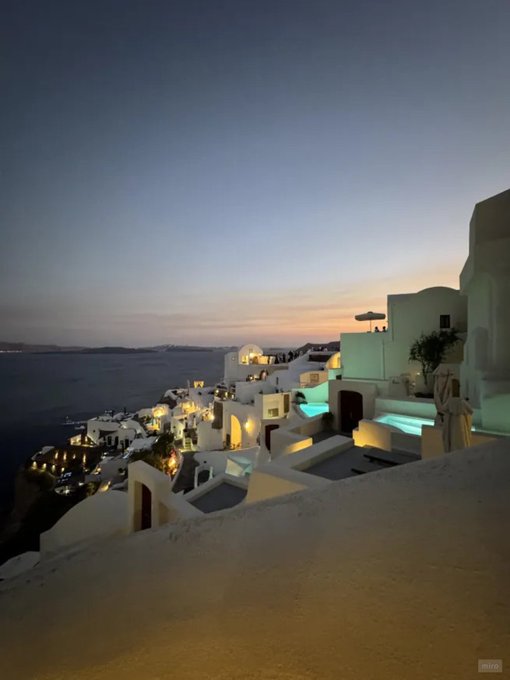 White-washed buildings of Santorini cascade down a cliffside at dusk, with pools glowing turquoise against the twilight sky. The serene Mediterranean scene suggests a dreamy, aspirational vibe fitting the optimistic "gm" greeting and excitement over upcoming NFT drops.