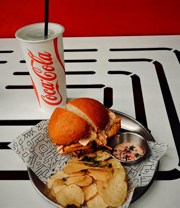 A meal served on a metal tray with patterned paper, featuring a chicken sandwich on a brioche bun, potato chips, and a small cup of pink dipping sauce, accompanied by a Coca-Cola cup with a straw on a white slatted table.