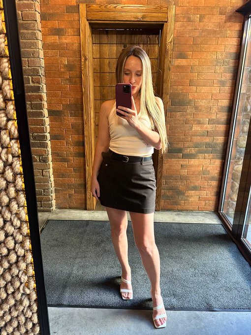 A mirror selfie of a woman with long blonde hair wearing a white sleeveless top, a black mini skirt, and white heeled sandals, standing in front of a brick wall.