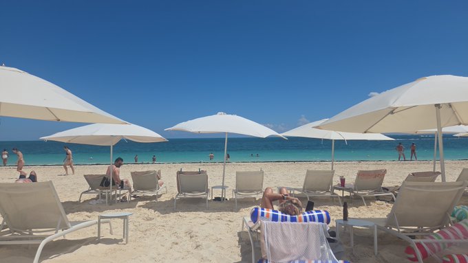 A bright sunny beach scene features white umbrellas, lounge chairs, and people relaxing on white sand against turquoise water under a clear blue sky.