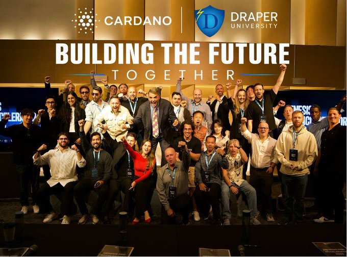 A group of enthusiastic participants celebrates together on stage with raised fists in front of a banner displaying "CARDANO" and "DRAPER UNIVERSITY" logos alongside the slogan "BUILDING THE FUTURE TOGETHER." The image visually captures the triumphant spirit of the Draper University Genesis program's collaboration with Cardano, reflecting the post's excitement about the program's impact and anticipation for the next APEX group.