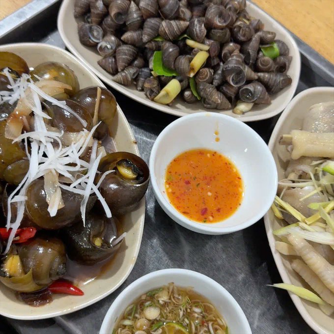 A tray of food featuring a large plate of cooked snails with shredded ginger on top, a plate of smaller snails, a plate of chicken feet, a dipping sauce, and a small bowl of noodle soup.
