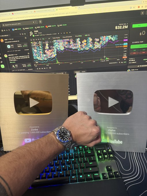 A photo shows a fist with a luxury watch resting on an RGB mechanical keyboard, with two YouTube Creator Awards (gold for 1,000,000 subscribers and silver for 100,000 subscribers) displayed in front of a monitor showing cryptocurrency charts with a market cap of $32.2M.