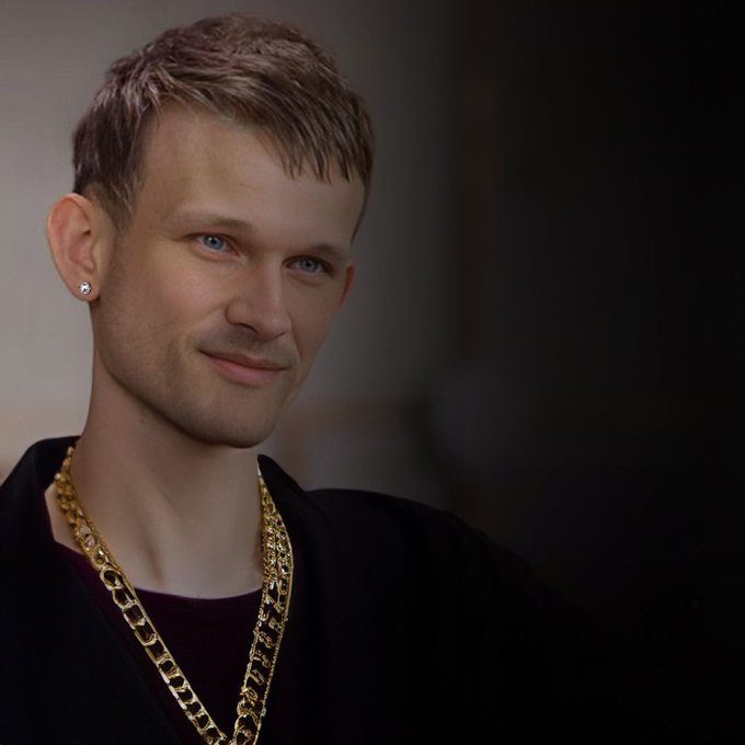 A man with bleached blond hair, blue eyes, and diamond stud earrings smiles slightly while wearing a black outfit layered with chunky gold chain necklaces, fitting the bold "quant" style referenced in the post.