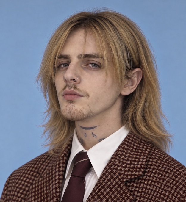 A close-up portrait shows a young man with shoulder-length blonde hair, a nose ring, facial piercings, neck tattoos, and a brown checkered suit with a dark tie against a blue background.