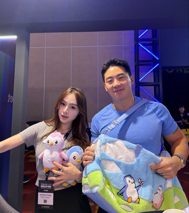 A woman holding two penguin plushies and a man in a blue shirt posing together with a blue fuzzy blanket, wearing a 'TOKEN2049 SINGAPORE' attendee badge.