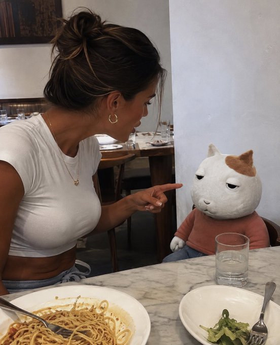 A woman in a white t-shirt points accusingly at a plush cat toy sitting at a restaurant table, with plates of pasta and greens in the foreground.