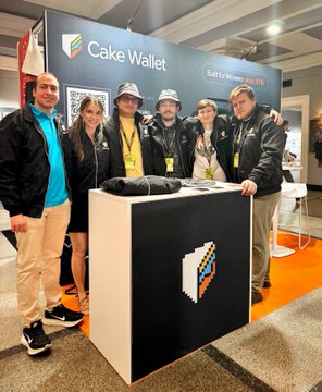 In the image, a group of six people are standing around a table with a sign that reads "The Cake Wallet". The individuals appear to be posing for a photo or attending an event related to cryptocurrency. One person is wearing a hat, while another has on a jacket. A man in the center of the group is holding a cell phone and appears to be taking a picture. The rest of the group is standing around the table, with one person on each side.