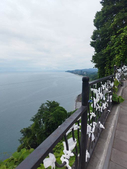 A coastal overlook featuring a black wrought-iron railing decorated with white ribbons, with a view of the ocean and distant shoreline under a cloudy sky.