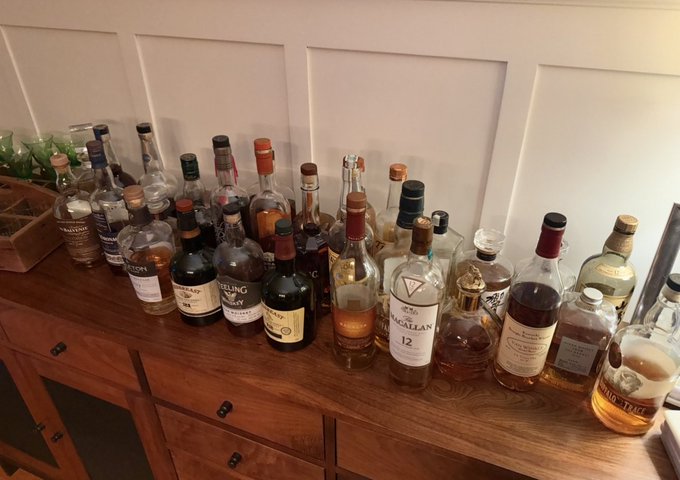 A wooden cabinet top lined with numerous partially empty liquor bottles, including brands like The Macallan 12.