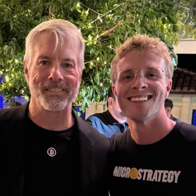 Two men smile for a photo at what appears to be a social event with greenery in the background; the younger man wears a black "MICROSTRATEGY" t-shirt (with the "O" styled as a Bitcoin logo), while the older man wears a black jacket featuring a Bitcoin pin. The image relates to the post as a nostalgic throwback featuring someone associated with early $MSTR enthusiasm being asked about.