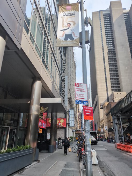 A street-level photograph of the Paramount Hotel entrance in New York City, showing a 'The Book of Mormon' theatrical banner hanging overhead and a commercial vehicle loading zone sign with zone number 102623.
