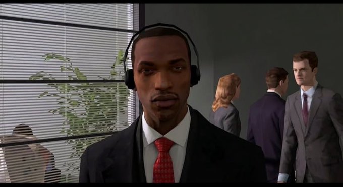 A screenshot from a video game showing a man in a suit and red tie wearing headphones in an office setting with other suited characters behind him.