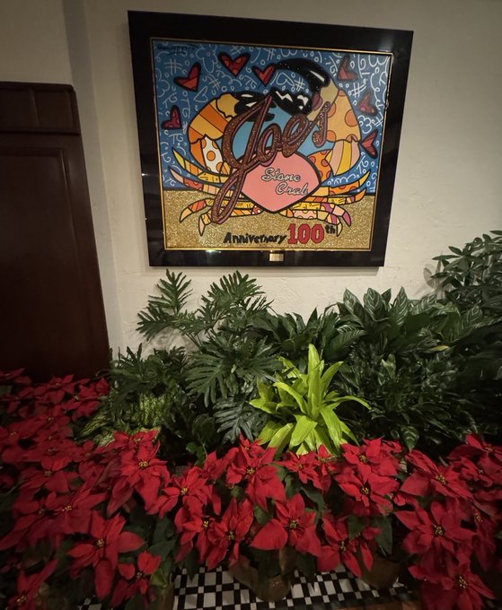 A framed colorful artwork of Joe's Stone Crab celebrating its 100th Anniversary hangs on a wall above a decorative arrangement of green plants and red poinsettias.