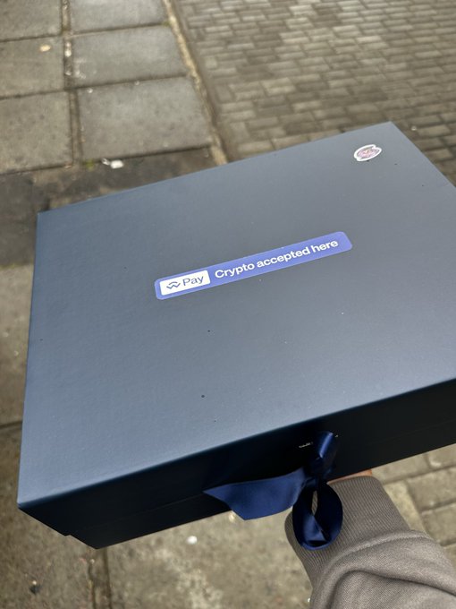A person holding a dark blue gift box with a navy ribbon outdoors on a brick sidewalk, featuring a sticker that reads 'Crypto accepted here' with a Pay logo.