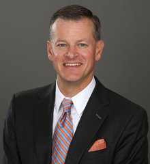 A professional headshot of a smiling man with short graying hair, wearing a black suit, light blue shirt, and a striped tie in orange, blue, and white, with a coordinating pocket square—colors associated with the University of Florida Gators, fitting the post about Florida's Director of Athletics welcoming Ripple ($XRP) to the Gator Nation.