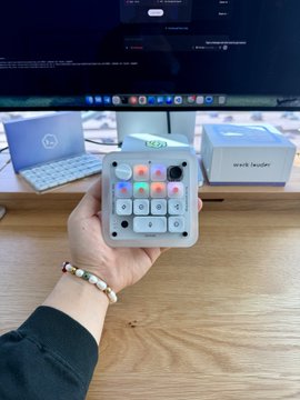 A hand holds a small black device called "work leader" up to the camera, showcasing its colorful illuminated buttons and control panel in front of a computer monitor. The compact hardware aligns with the caption about setting up a new device.