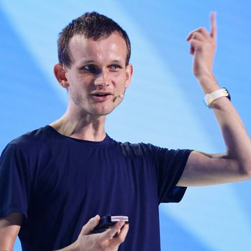 Vitalik Buterin speaks on stage gesturing with his right hand, wearing a navy blue t-shirt and a white watch, while holding a small device. His pointed pose complements the post's bold quote about Ethereum's evolving role and hints at his confident outlook on the blockchain's future growth.