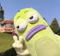 A bright yellow inflatable mascot character with large eyes, a purple mustache, and a peace sign gesture standing on grass at what appears to be a theme park.
