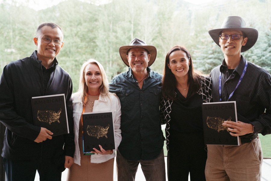 A group photo of five people posing together while three of them hold up copies of a book titled 'AMERICAN' featuring a US map on the cover, with mountain scenery in the background.