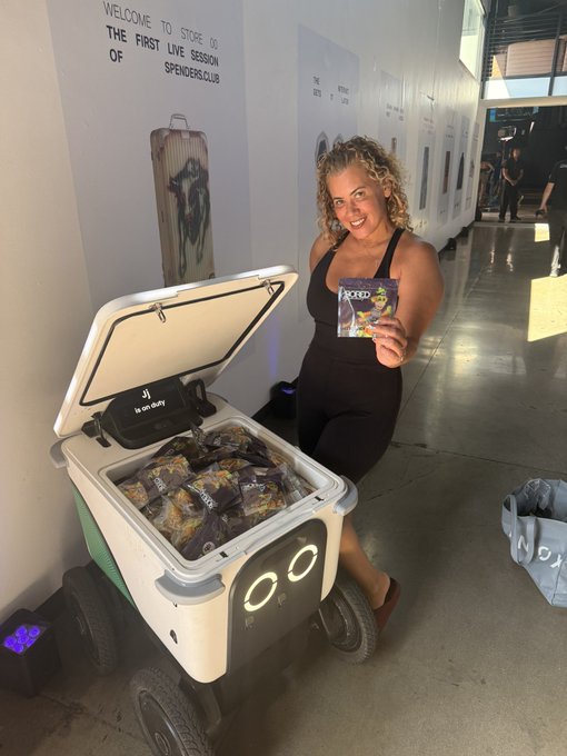 A woman at 'SPENDERS CLUB' store smiling while holding a booklet, standing next to a white cooler on wheels filled with packaged snacks.