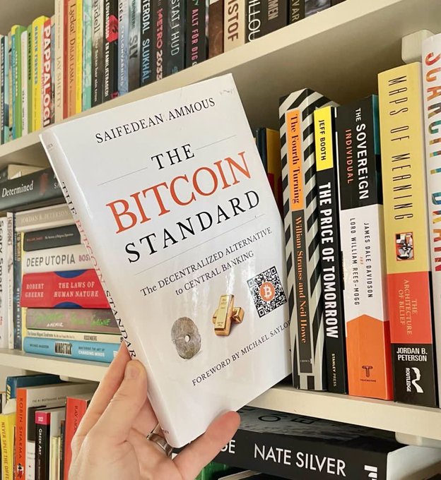 A hand holding up the book "The Bitcoin Standard: The Decentralized Alternative to Central Banking" by Saifedean Ammous, photographed against a background bookshelf featuring titles like "Maps of Meaning" and "The Price of Tomorrow."