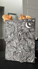 An ornate journal or book with an embossed silver cover depicting flowing waves, flowers, and a crescent moon, accented with dried orange flowers peeking from the top.