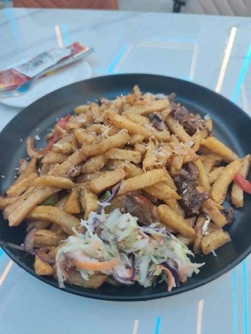 A black plate filled with french fries mixed with meat, topped with grated cheese and accompanied by a side of coleslaw with carrots and red cabbage.
