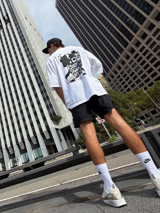 A low-angle streetwear photo of a person wearing a white graphic t-shirt, black shorts, white Nike socks, and sneakers, standing against a backdrop of tall city skyscrapers.