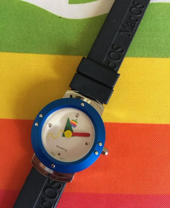 A vintage MacOS Quartz watch with a blue bezel and black rubber band resting on a striped green, yellow, and red surface.