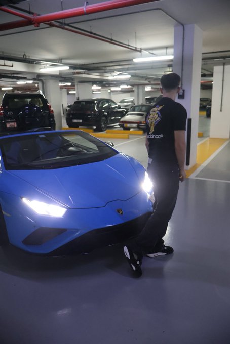 A man wearing a black t-shirt featuring a Lamborghini logo stands next to a striking blue Lamborghini Huracán with its headlights illuminated in an underground parking garage. The "GN" post likely refers to the initials or social media handle of the owner posing proudly with their luxury supercar.