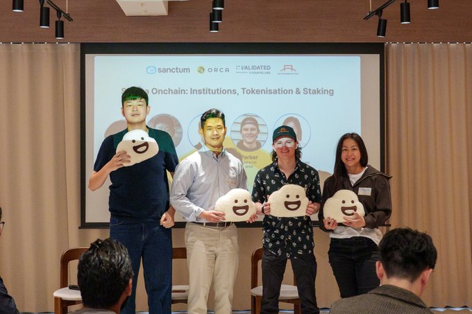 Four people stand on stage holding white smiley face cutouts in front of a projection screen reading 'sanctum ORCA VALIDATED' and 'Onchain: Institutions, Tokenisation & Staking'.