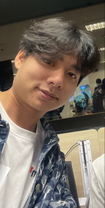 A young man with dark hair takes a selfie wearing a white t-shirt under a blue denim jacket, with a computer monitor visible in the background.