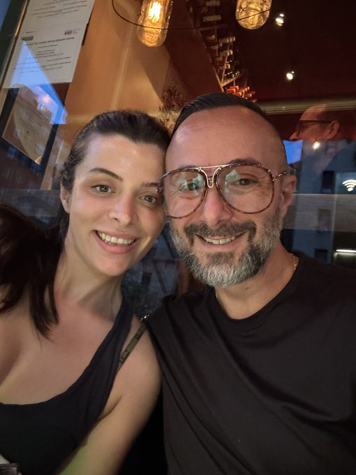 A selfie of a smiling couple—a woman with dark hair in a black tank top and a bearded man wearing glasses and a black shirt—posing together in what appears to be a dimly lit restaurant.