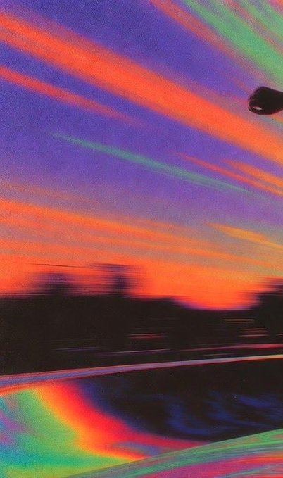 A motion-blurred photograph taken from a moving vehicle, capturing a vibrant sunset with streaks of orange, green, and rainbow-like light trails across the sky.