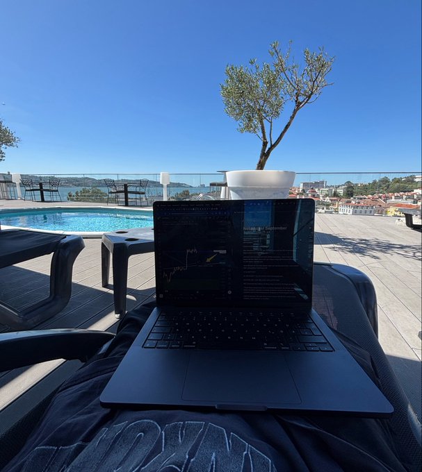 A laptop displaying a trading chart and text sits on a lounge chair by a rooftop pool overlooking a coastal town with a small tree and blue sky.