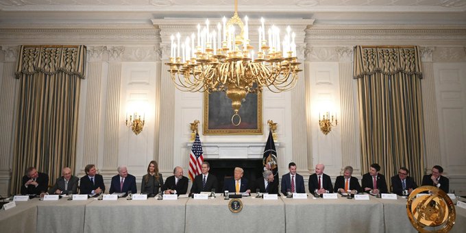 President Trump sits at the center of a long table in the White House's State Dining Room, flanked by officials and executives—including a gold Bitcoin visible in the foreground—gathering for the administration's largest-ever crypto policy meeting focused on regulation and the CLARITY Act.