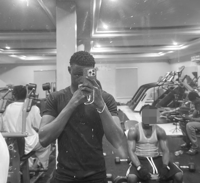 A black-and-white mirror selfie of a muscular man in a dark t-shirt taking a photo with his phone in a gym, with another person visible in the background whose face is blurred.