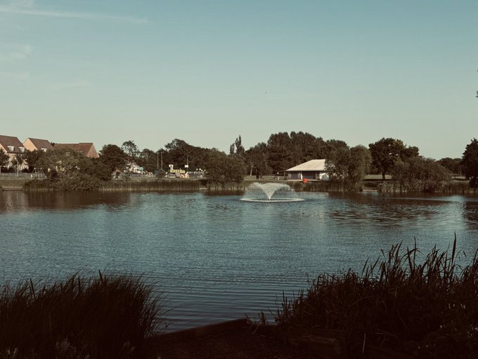 A serene landscape featuring a pond with a central water fountain, bordered by reeds and trees, with houses and a small building visible in the background under a clear blue sky.
