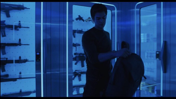 A man in a dark shirt standing in a blue-lit armory, handling a bag in front of glass display cases filled with various firearms.